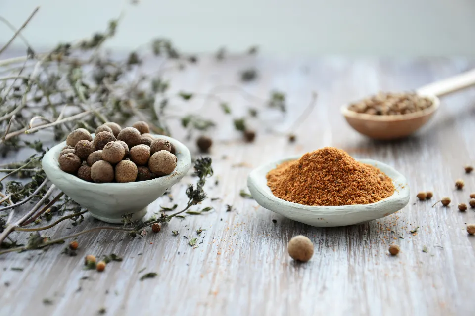 Whole dried spice berries in a white dish beside ground spice on a wooden table